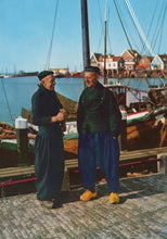 Load image into Gallery viewer, Two men standing on a dock with boats and a village in the background