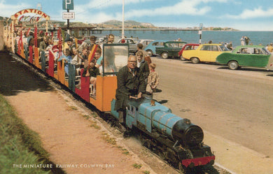 Miniature train at Colwyn Bay with people and vintage cars in the background.
