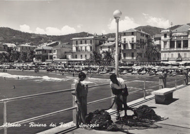 Two people standing on a promenade by a beach with buildings and water in the background.