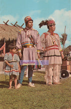 Load image into Gallery viewer, Three people in traditional Native American attire standing outdoors with a thatched structure and trees in the background.
