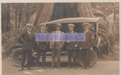 Group of men in formal attire standing next to an old-fashioned car in a forest setting.