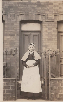 Woman in a white apron standing in front of a door with a brick wall background