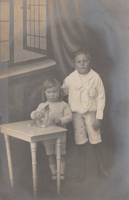 Two children standing near a small table with a glass object, in a vintage indoor setting.