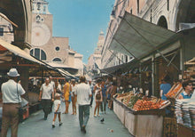 Load image into Gallery viewer, People walking through a market with fruit stands and architectural buildings in the background.
