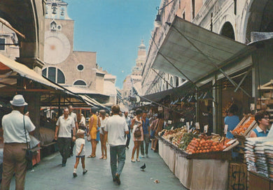 People walking through a market with fruit stands and architectural buildings in the background.