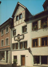 Load image into Gallery viewer, Traditional European building with wooden shutters and a decorative plaque on a street.
