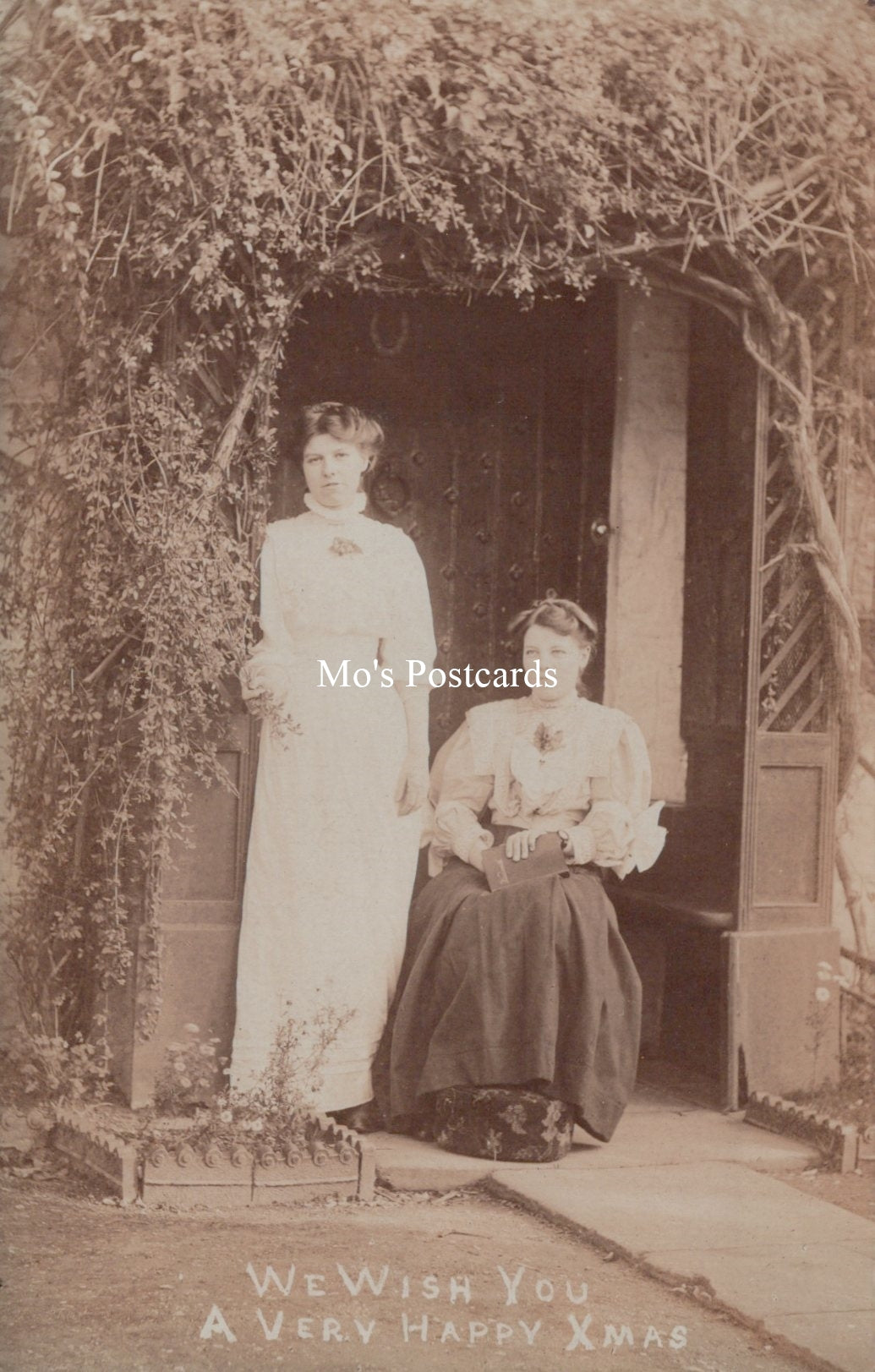 Vintage photograph of two women standing under a tree with a door in the background, labeled 'Mo's Postcards'.