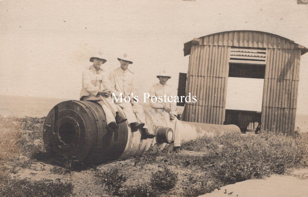 Vintage black and white photo of three men sitting on a large cylindrical object with a corrugated metal shed in the background.