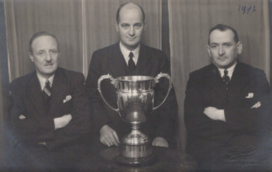Three men in suits posing with a large trophy in front of a curtain, with '1942' written on it.