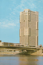 Load image into Gallery viewer, Tall beige building with a blue sky and water reflection
